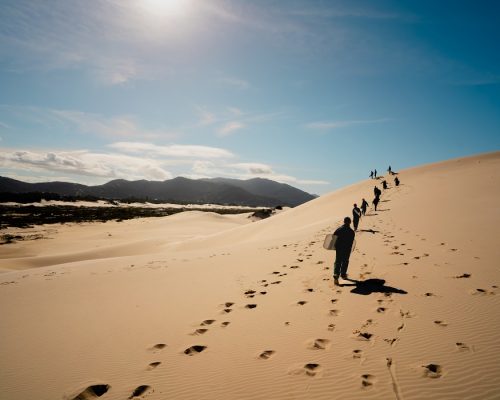 Areal nas dunas de Florianópolis; pessoas em fila a andar no deserto a deixar pegadas na areia